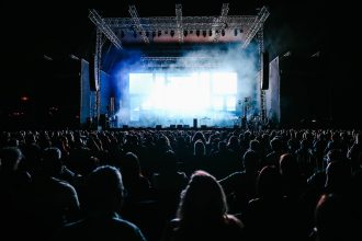 Grande público acompanha um show ao vivo em um festival de música ao ar livre, sob luzes coloridas.
