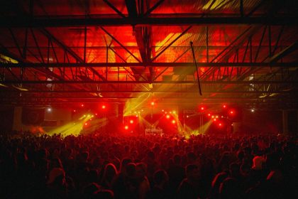 Público acompanha um show em ambiente fechado, sob iluminação vermelha e amarela.