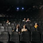Público diversificado sentado em uma sala de cinema pouco iluminada durante uma sessão
